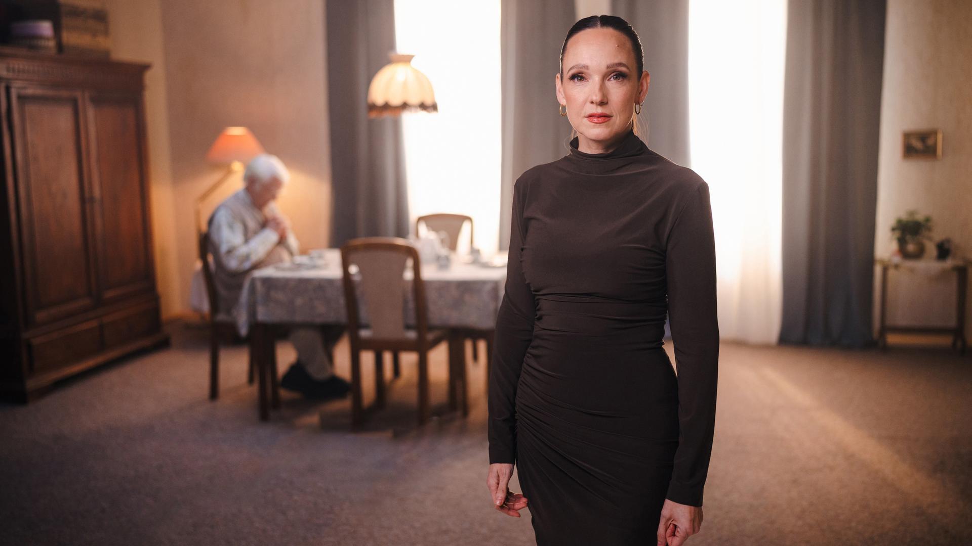Komikerin Carolin Kebekus schaut ernst in Richtung der Kamera. Im Hintergrund sitzt ein älterer Mensch einsam an einem großen Tisch. Komikerin Carolin Kebekus schaut ernst in Richtung der Kamera. Im Hintergrund sitzt ein älterer Mensch einsam an einem großen Tisch.