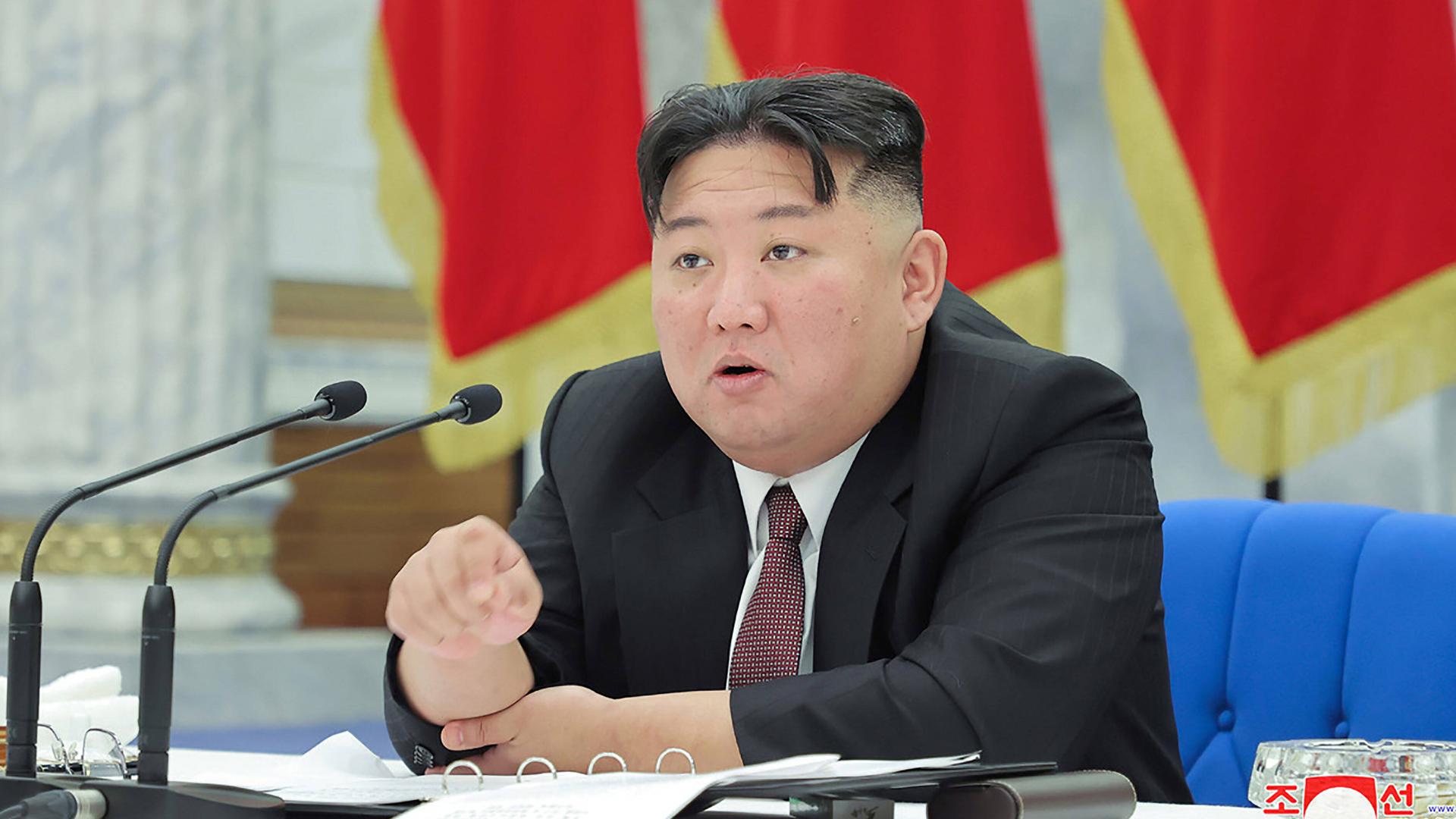 Das Bild zeigt den nordkoreanischen Machthaber Kim Jong-un sitzend während eines Vortrags. Im Hintergrund sind Flaggen zu sehen.