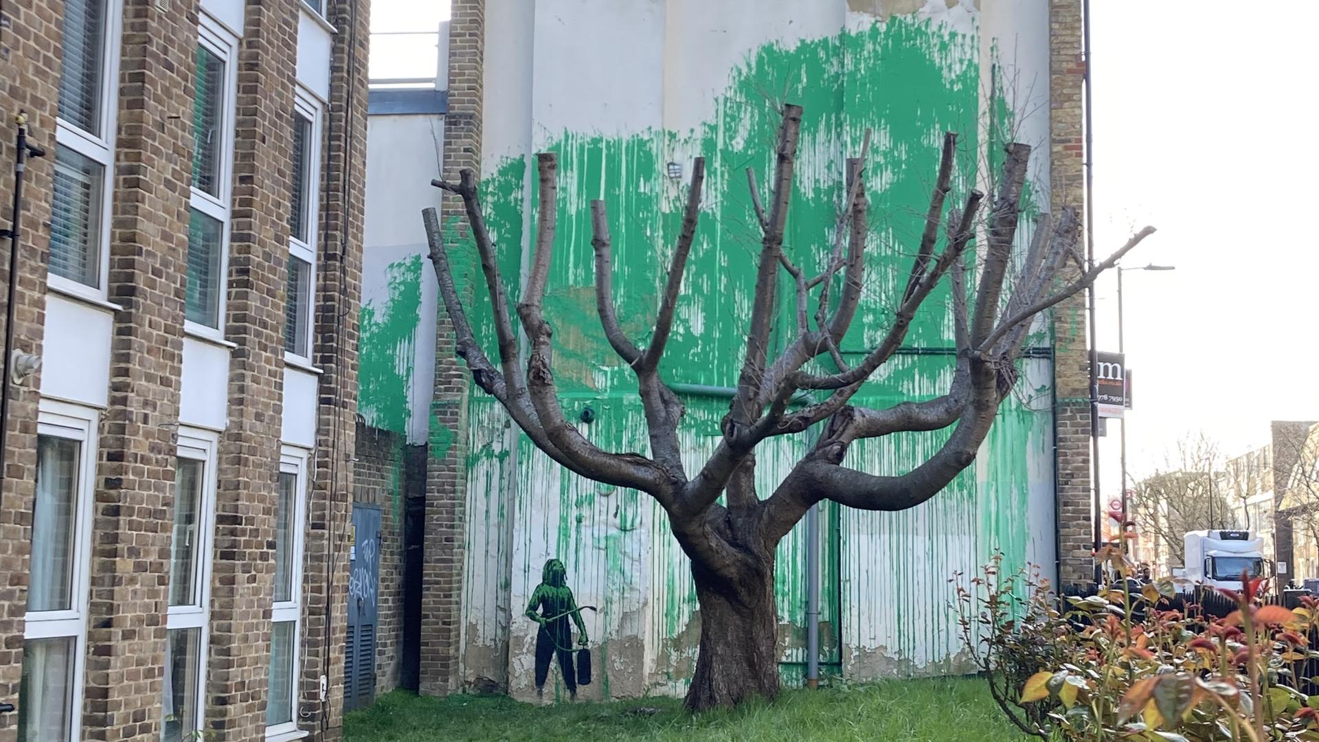 Auf eine Hauswand ist ein Bild gemalt. Es vermittelt den Eindruck, als würde der reale Baum davor Blätter tragen.