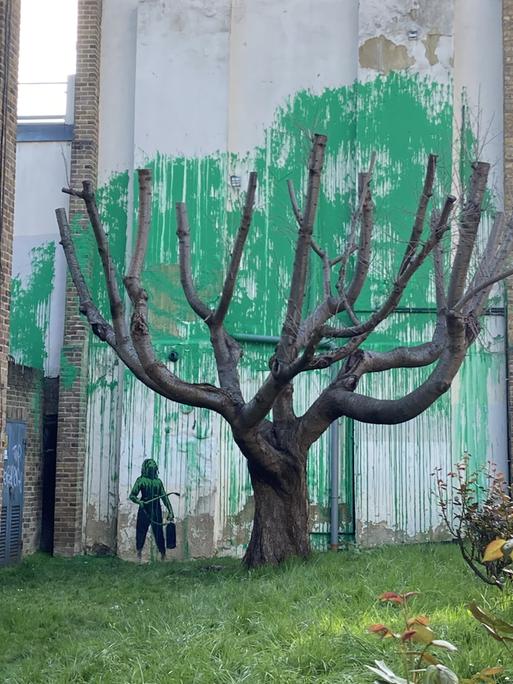Auf eine Hauswand ist ein Bild gemalt. Es vermittelt den Eindruck, als würde der reale Baum davor Blätter tragen.