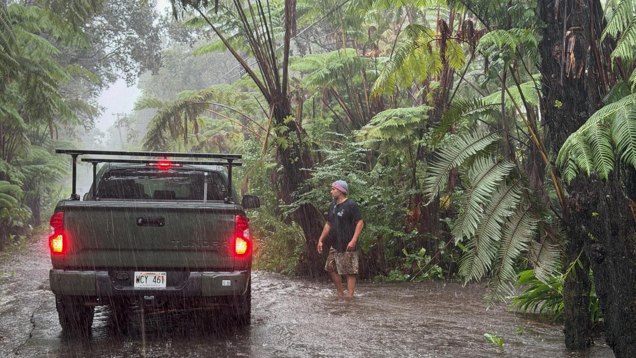 Ein Pickup steht auf einer Straße in einer regenwaldähnlichen Vegetation im starken Regen. Ein Mann mit Mütze geht auf das Fahrzeug zu. 