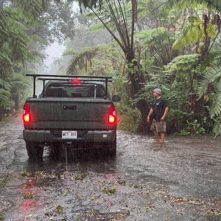 Ein Pickup steht auf einer Straße in einer regenwaldähnlichen Vegetation im starken Regen. Ein mann mit Mütze geht auf das Fahrzeug zu. 