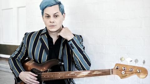 Jack White ist von der Hüfte an aufwärts zu sehen. Er hat die Haare hellblau gefärbt, sein Sakko ist in hellblau und  schwarz länggestreift, darunter trägt ein in dem hellen Raum ein schwarzes Oberteil. Vor dem Bauch hält er eine Bassgitarre.