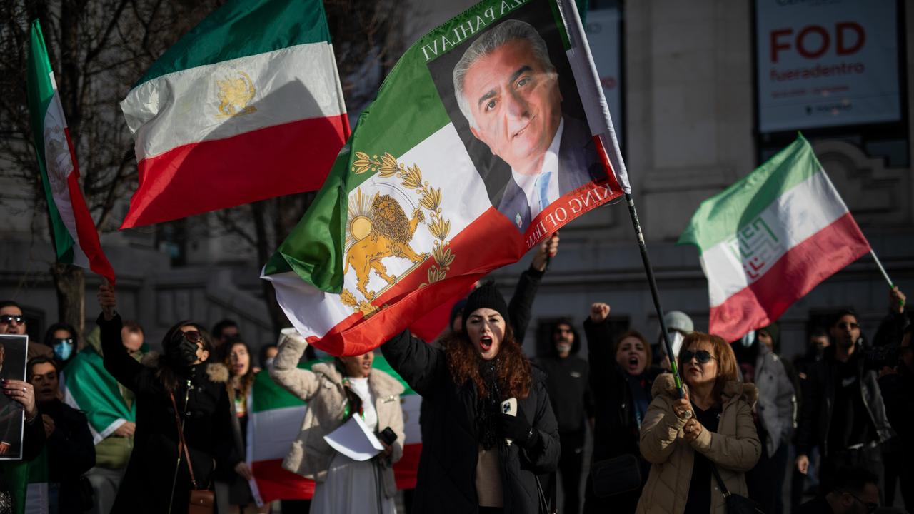 Mehrere Menschen halten grün-weiß-rote iranische Flaggen ohne das Emblem der Islamischen Republik hoch. In der Bildmitte trägt eine Person eine Fahne mit einem Porträt von Reza Pahlavi, dem Sohn des 1979 gestürzten Schahs. Mehrere Menschen halten grün-weiß-rote iranische Flaggen ohne das Emblem der Islamischen Republik hoch. In der Bildmitte trägt eine Person eine Fahne mit einem Porträt von Reza Pahlavi, dem Sohn des 1979 gestürzten Schahs.