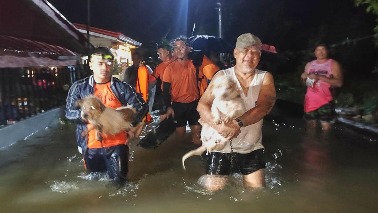Es ist dunkel. Mehrere Menschen, darunter Rettungskräfte in orangefarbenen Warnwesten, gehen eine überflutete beleuchtete Straße entlang. Sie stehen knietief im Wasser. Ein Mann trägt einen Hund. 