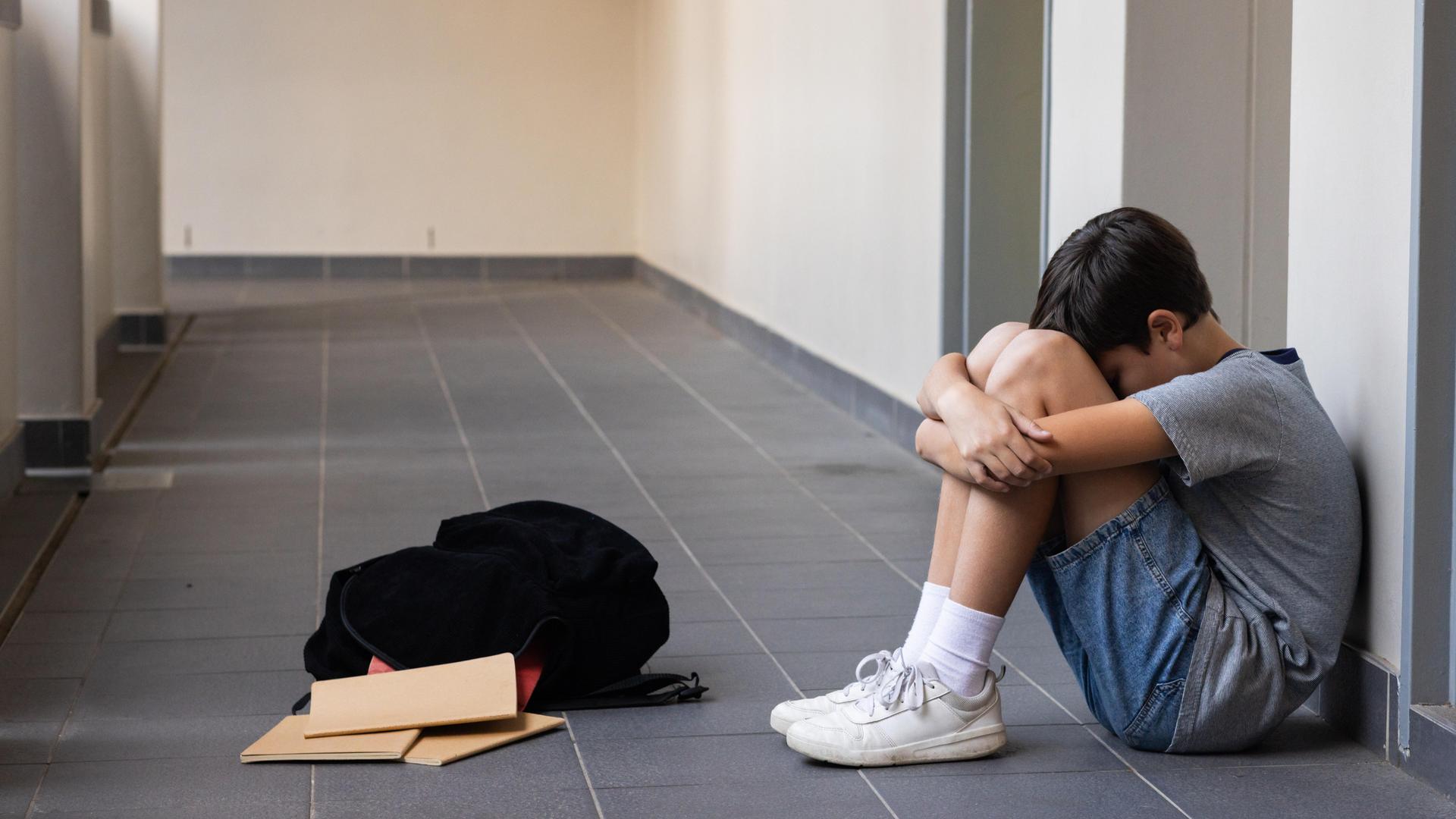 Eine Junge sitzt auf dem Boden und umarmt seine Knie. Vor ihm liegen sein Rucksack und Schulhäfte. Eine Junge sitzt auf dem Boden und umarmt seine Knie. Vor ihm liegen sein Rucksack und Schulhäfte.
