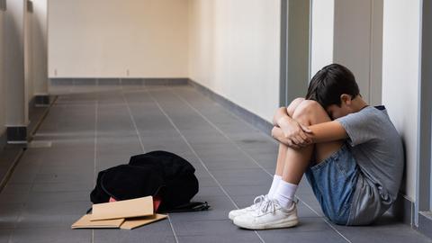 Eine Junge sitzt auf dem Boden und umarmt seine Knie. Vor ihm liegen sein Rucksack und Schulhäfte. 