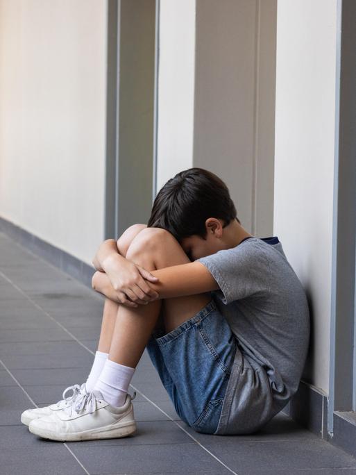 Eine Junge sitzt auf dem Boden und umarmt seine Knie. Vor ihm liegen sein Rucksack und Schulhäfte. 