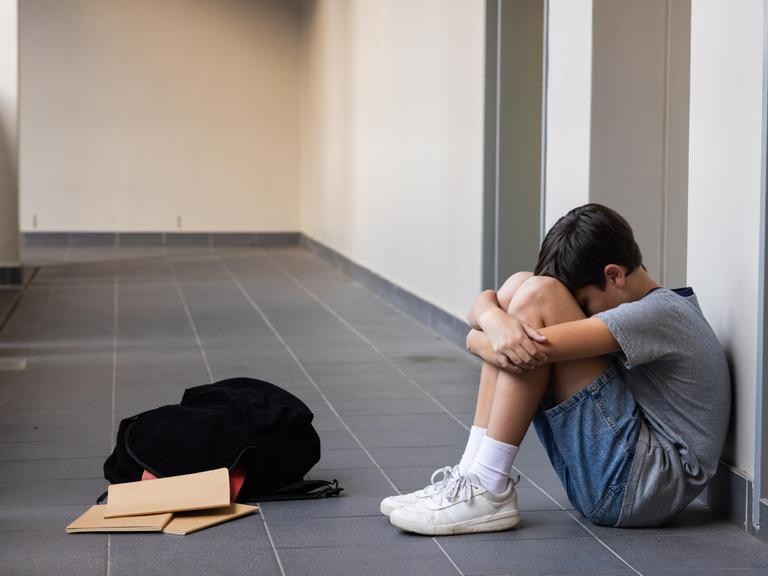 Eine Junge sitzt auf dem Boden und umarmt seine Knie. Vor ihm liegen sein Rucksack und Schulhäfte. 