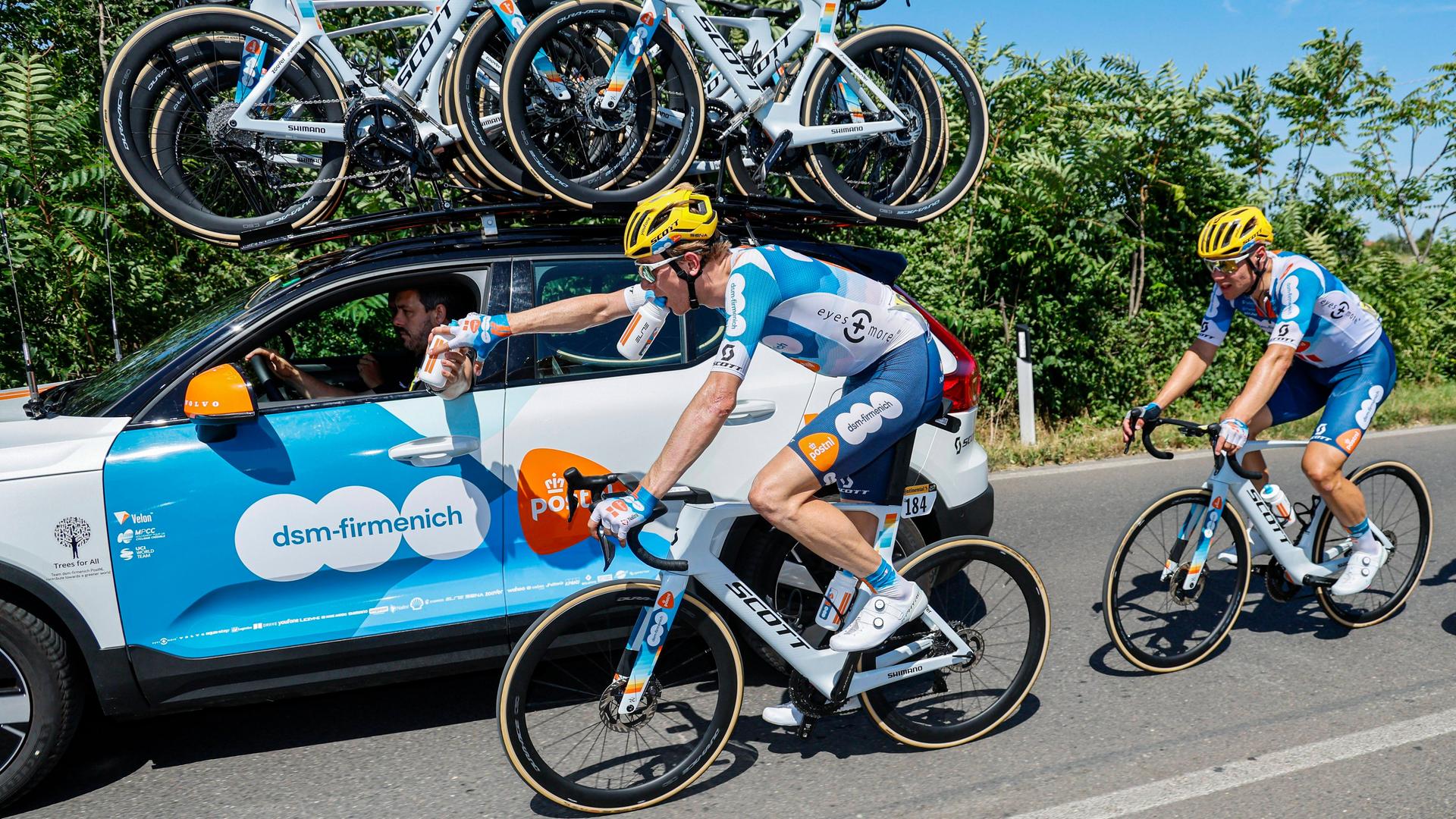 Ein Mannschaftswagen fährt bei der Tour de France neben Nils Eekhoff (dsm-firmenich PostNL), ihm wird eine Trinkflasche gereicht.