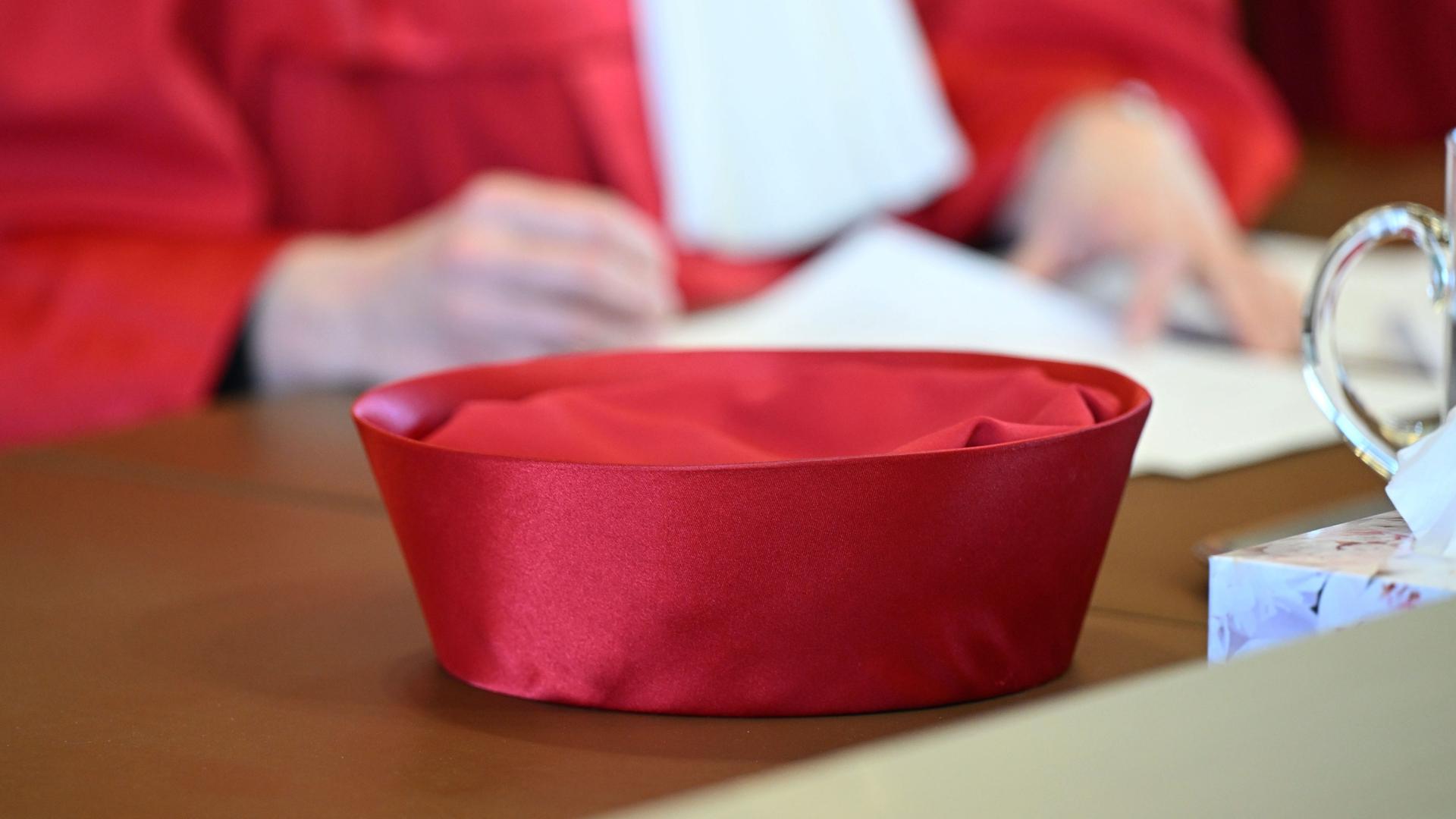 Zu sehen ist die roten Robe einer Bundesverfassungsrichterin, im Vordergrund sieht man den Hut auf dem Tisch liegen, im Hintergrund sieht man in der Unschärfe zwei Hände, die ein Papier halten.