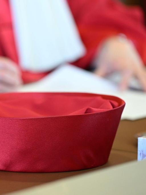 Zu sehen ist die roten Robe einer Bundesverfassungsrichterin, im Vordergrund sieht man den Hut auf dem Tisch liegen, im Hintergrund sieht man in der Unschärfe zwei Hände, die ein Papier halten.