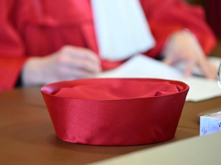 Zu sehen ist die roten Robe einer Bundesverfassungsrichterin, im Vordergrund sieht man den Hut auf dem Tisch liegen, im Hintergrund sieht man in der Unschärfe zwei Hände, die ein Papier halten.