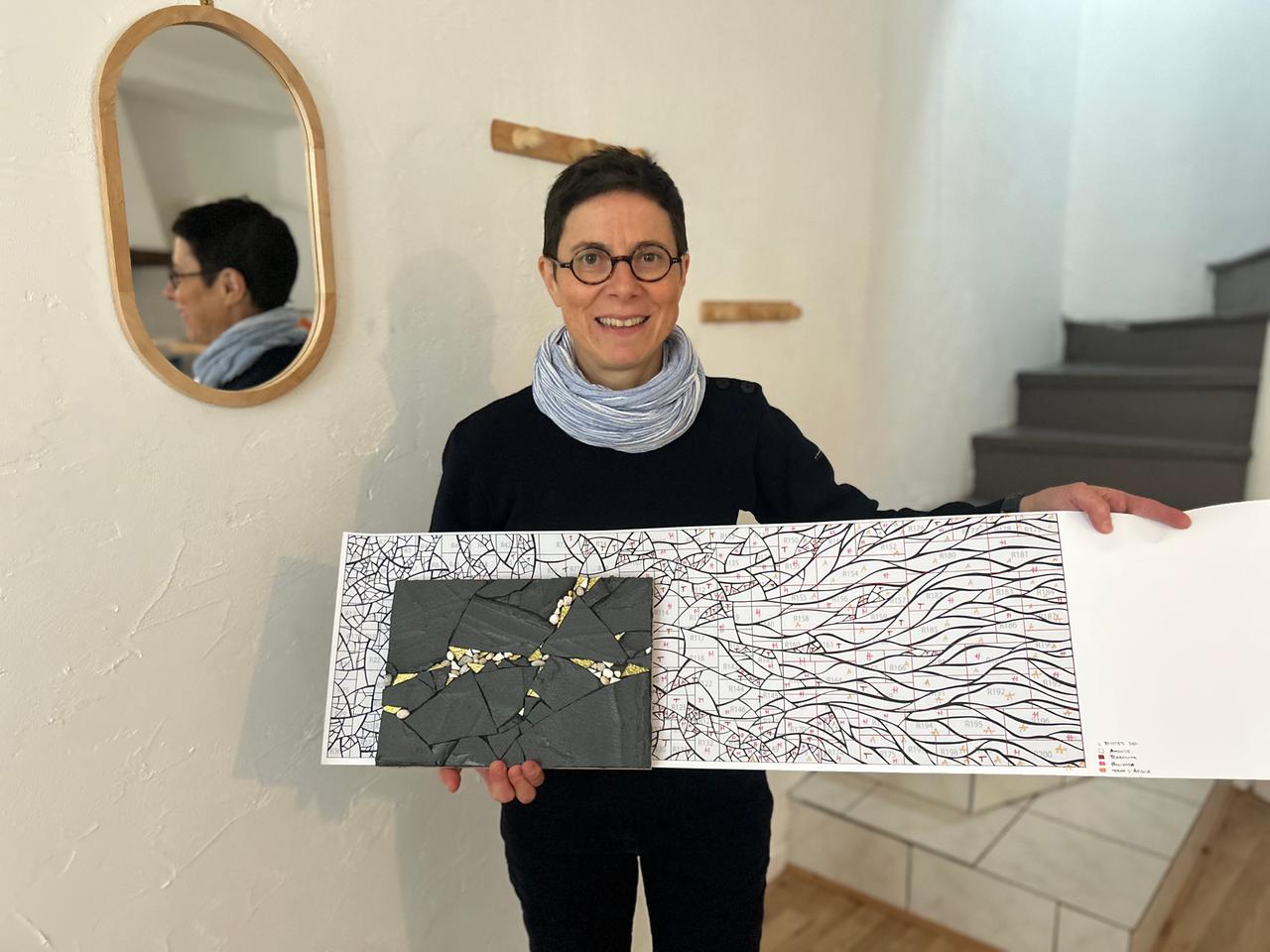 Schwester Samuelle, eine lächelnde Frau mit Brille und Kurzhaarfrisur, hält einen Entwurf zu einem Mosaik mit einigen schwarzen Mosaiksteinen in den Händen. Ihr Profil ist in einem Spiegel an der Wand zu erkennen.