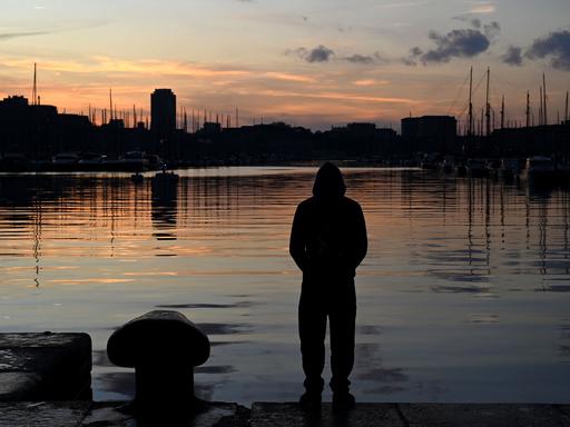Silhouette eines Mannes im Kapuzenpullover, der den Sonnenuntergang über dem Vieux-Port in Marseille betrachtet.