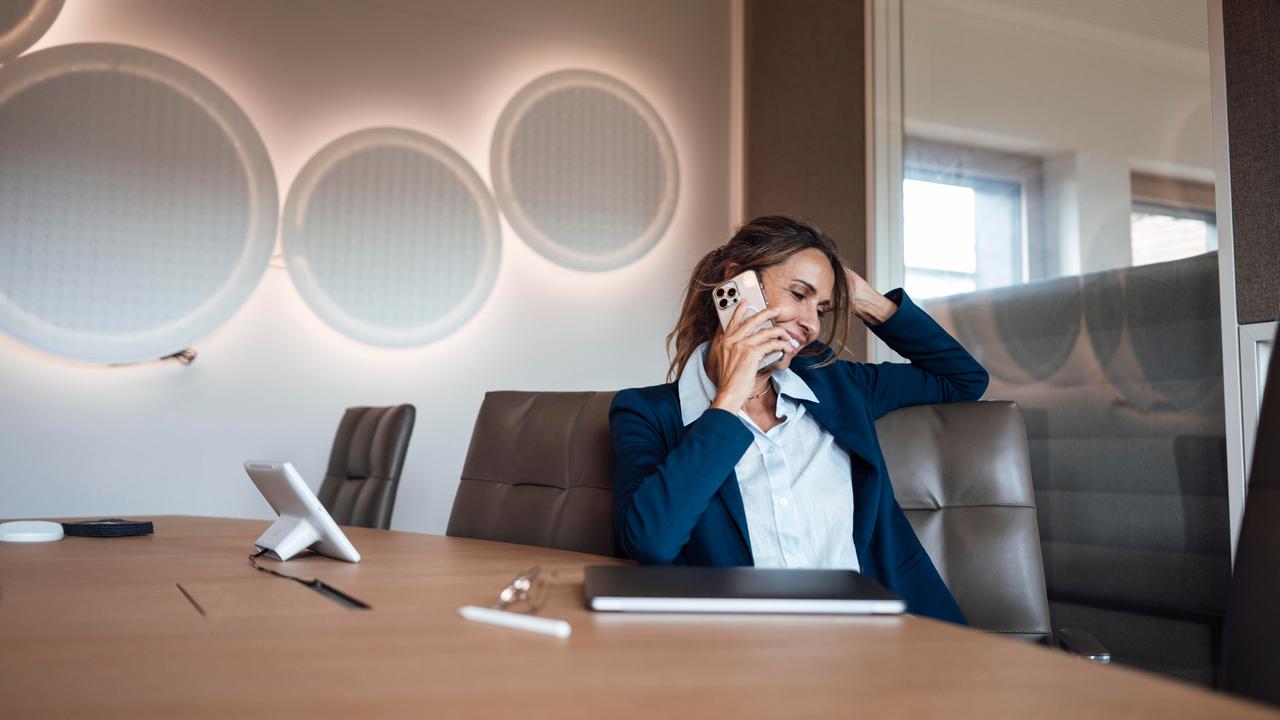 Die Frau im dunkelblauen Kostüm sitzt lächelnd an einem Konferenztisch und telefoniert. Dahinter eine stylische Wand mit kreisförmigen Lampen. 