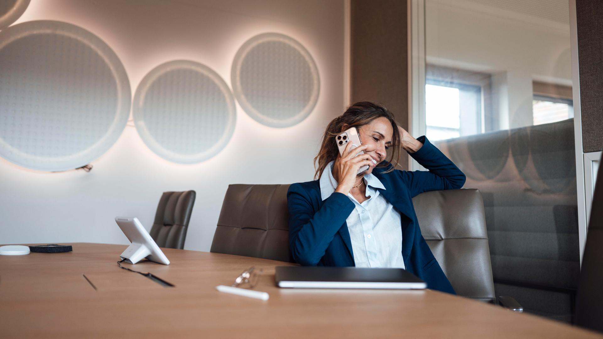 Die Frau im dunkelblauen Kostüm sitzt lächelnd an einem Konferenztisch und telefoniert. Dahinter eine stylische Wand mit kreisförmigen Lampen. Die Frau im dunkelblauen Kostüm sitzt lächelnd an einem Konferenztisch und telefoniert. Dahinter eine stylische Wand mit kreisförmigen Lampen.