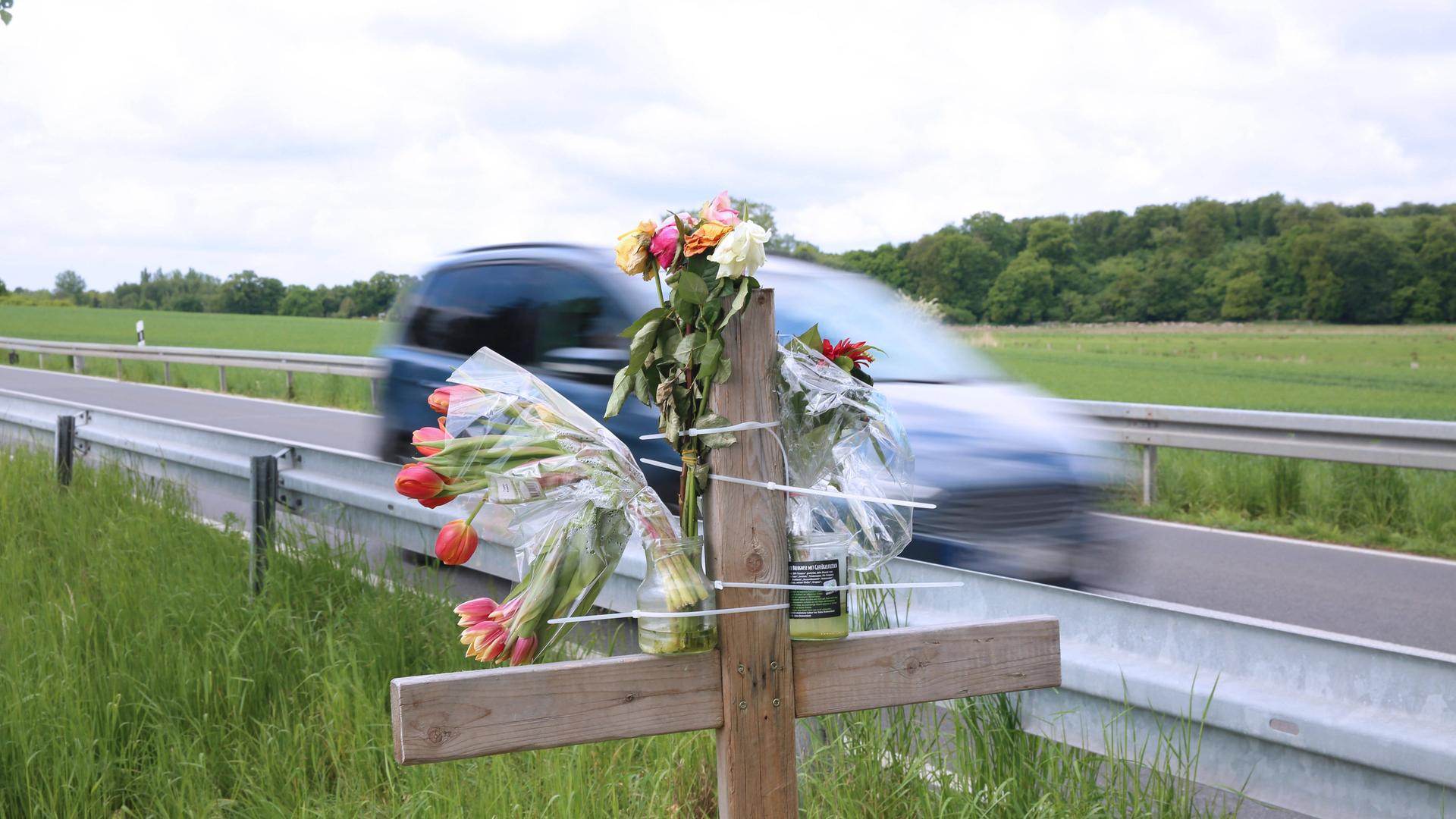 Am Straßenrand einer Landstraße stehen ein Kreuz und Blumen. Ein Auto fährt vorbei.