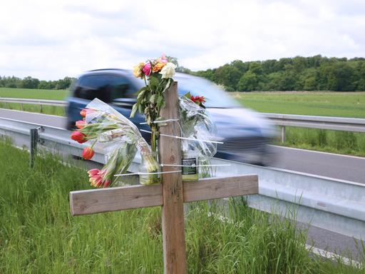 Am Straßenrand einer Landstraße stehen ein Kreuz und Blumen. Ein Auto fährt vorbei.