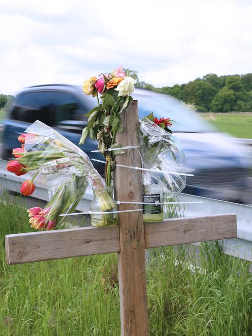 Am Straßenrand einer Landstraße stehen ein Kreuz und Blumen. Ein Auto fährt vorbei.