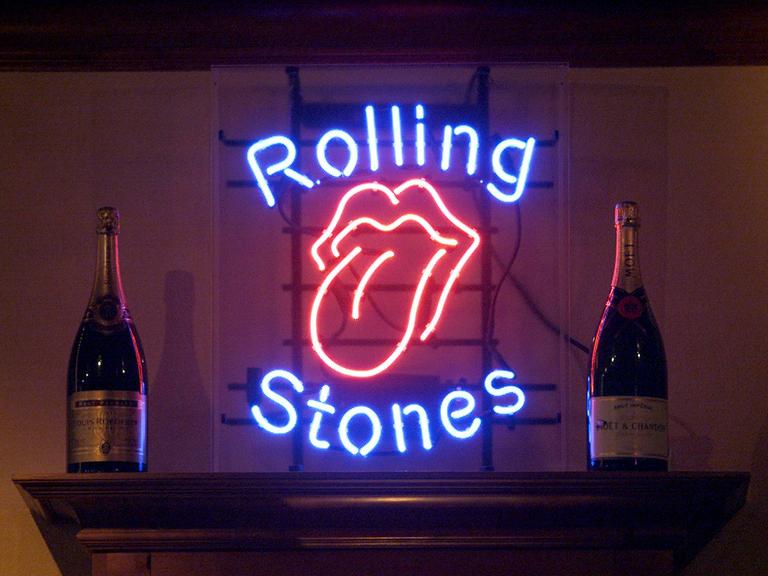 Eine Leuchtreklame mit typischen Rolling Stones Logo einer rausgestreckten Zunge und dem Schrift der Band flankiert von zwei Champagner-Flaschen.