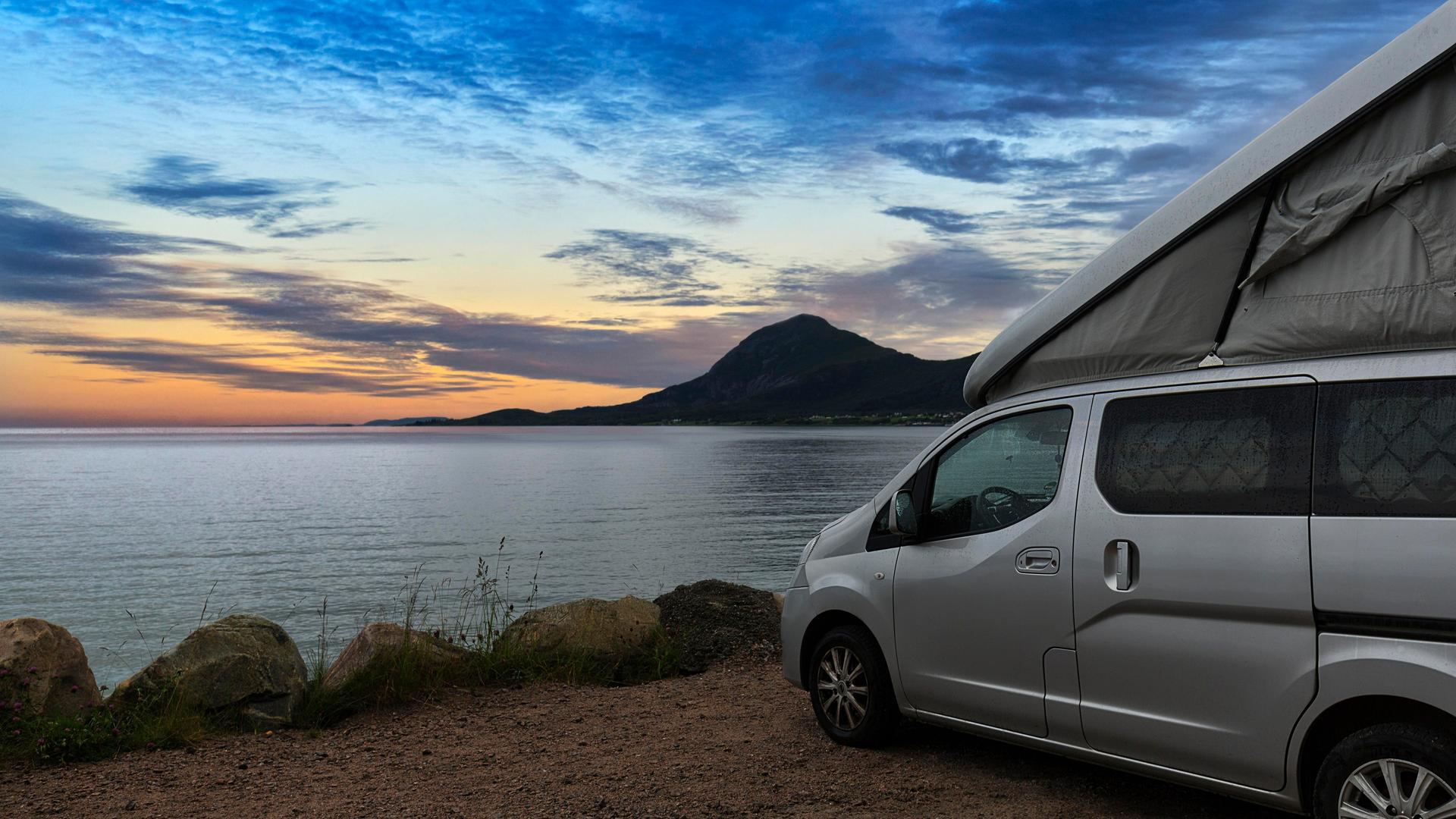 Ein Campervan mit Aufstelldach steht in der Abenddämmerung auf einem Parkplatz am Meer in Norwegen.