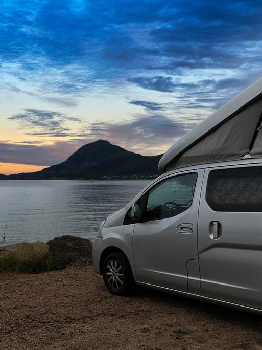 Ein Campervan mit Aufstelldach steht in der Abenddämmerung auf einem Parkplatz am Meer in Norwegen.