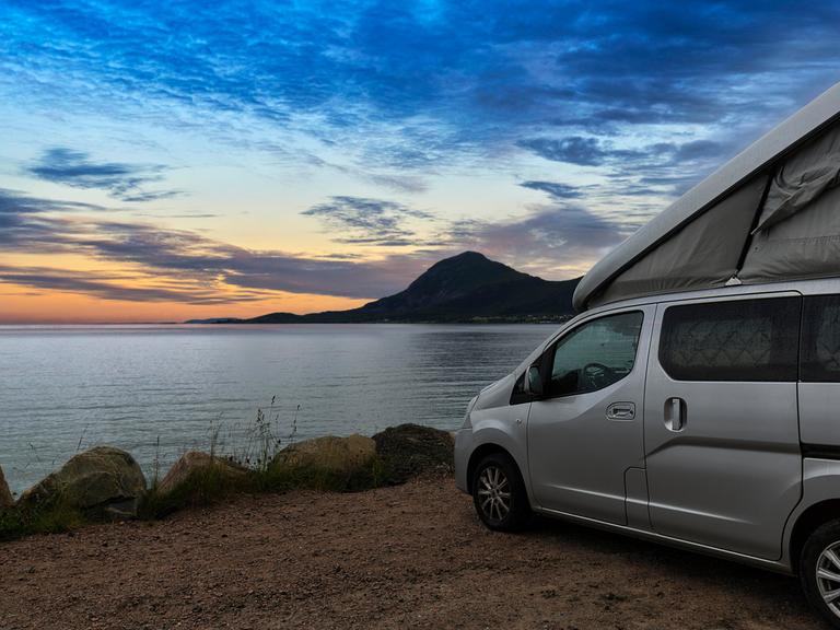 Ein Campervan mit Aufstelldach steht in der Abenddämmerung auf einem Parkplatz am Meer in Norwegen.