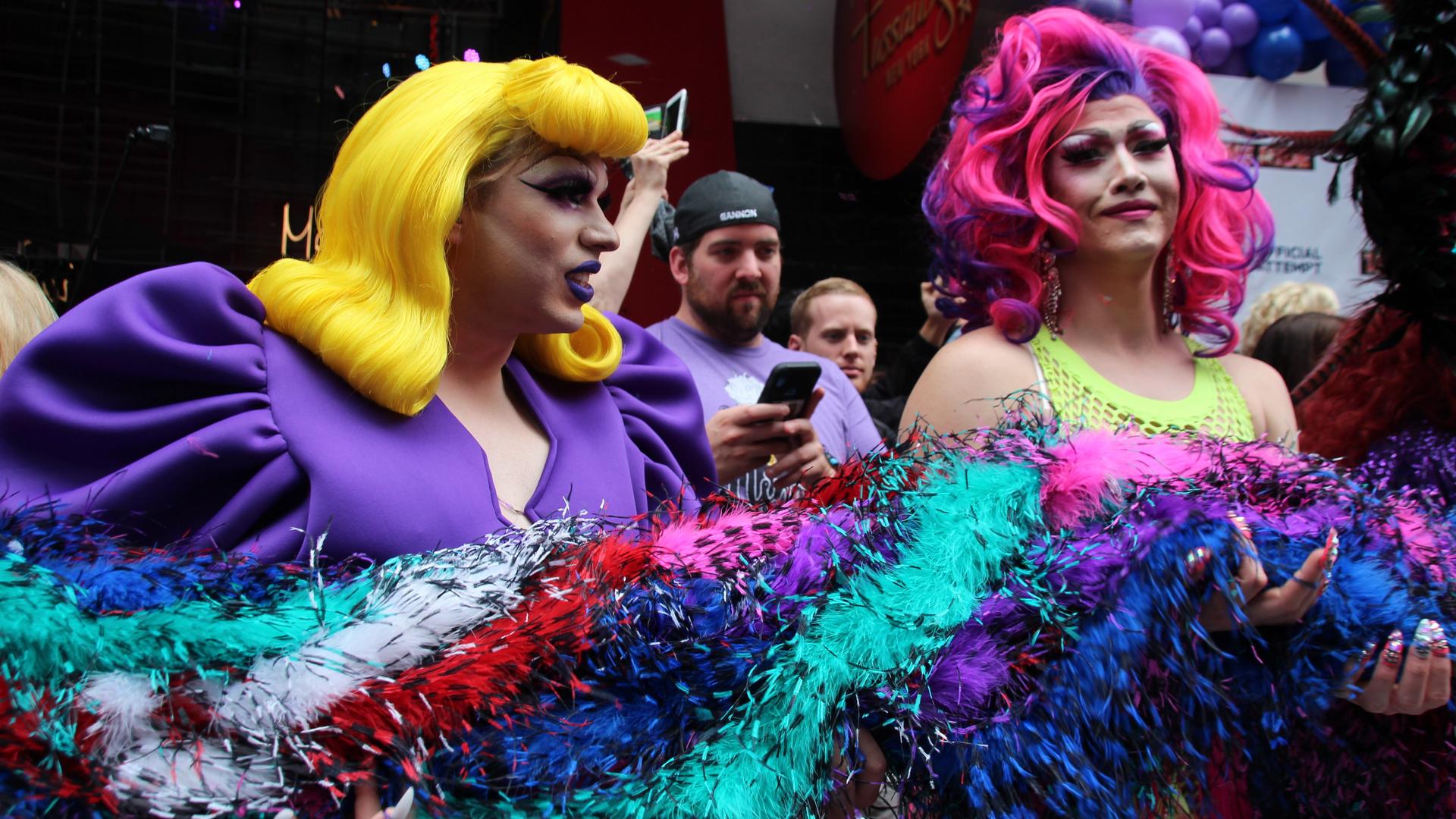 Zwei als Frauen gekleidete Mänder halten eine dicke bunte Federboa. Die eine Drag Queen hat eine lange gelbe Perrücke mit voluminösem Pony und ein lila farbenes Kleid mit dicken Puffärmeln an. Die andere Drag Queen trägt eine lila und pink farbenen Perrücke und ein neongelbes Netz-Oberteil. Beide sind stark geschminkt. Zwei als Frauen gekleidete Mänder halten eine dicke bunte Federboa. Die eine Drag Queen hat eine lange gelbe Perrücke mit voluminösem Pony und ein lila farbenes Kleid mit dicken Puffärmeln an. Die andere Drag Queen trägt eine lila und pink farbenen Perrücke und ein neongelbes Netz-Oberteil. Beide sind stark geschminkt.