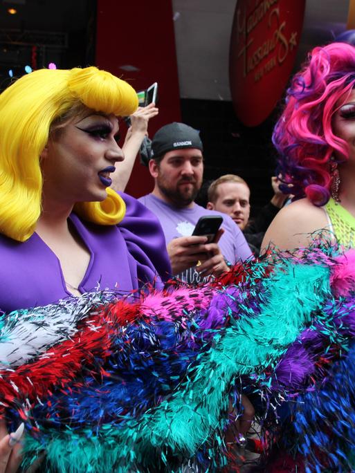 Zwei als Frauen gekleidete Mänder halten eine dicke bunte Federboa. Die eine Drag Queen hat eine lange gelbe Perrücke mit voluminösem Pony und ein lila farbenes Kleid mit dicken Puffärmeln an. Die andere Drag Queen trägt eine lila und pink farbenen Perrücke und ein neongelbes Netz-Oberteil. Beide sind stark geschminkt.