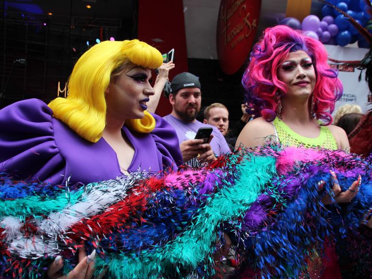 Zwei als Frauen gekleidete Mänder halten eine dicke bunte Federboa. Die eine Drag Queen hat eine lange gelbe Perrücke mit voluminösem Pony und ein lila farbenes Kleid mit dicken Puffärmeln an. Die andere Drag Queen trägt eine lila und pink farbenen Perrücke und ein neongelbes Netz-Oberteil. Beide sind stark geschminkt.