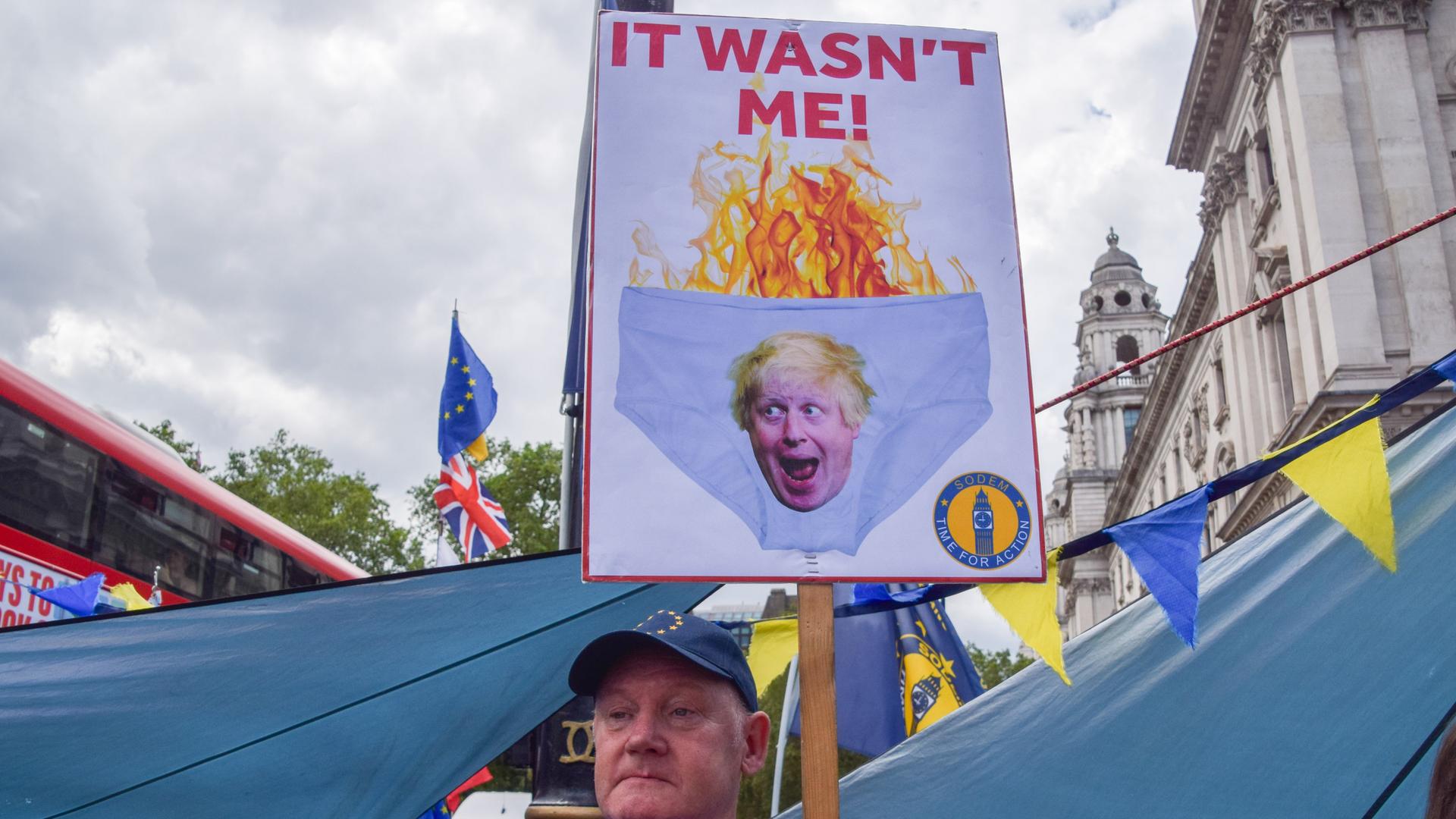 Ein Demonstrant hält ein Schild in die Luft, welches ein illustriertes Portrait von Boris Johnson auf einer brennenden Unterhose mit der Aufschrift "It wasn't me" zeigt
