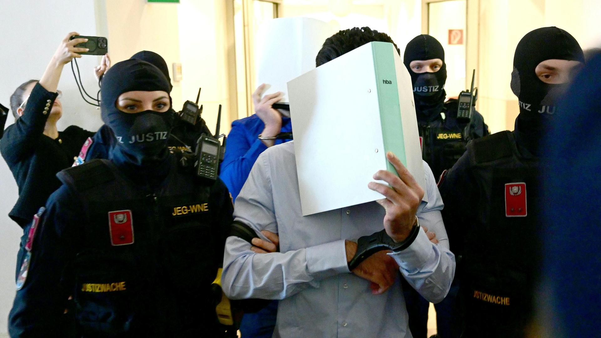 Polizisten führen zwei Männer in in einen Gerichts-Saal. Die Männer tragen Handschellen. Sie halten sich Aktenordner vor das Gesicht. Polizisten führen zwei Männer in in einen Gerichts-Saal. Die Männer tragen Handschellen. Sie halten sich Aktenordner vor das Gesicht.