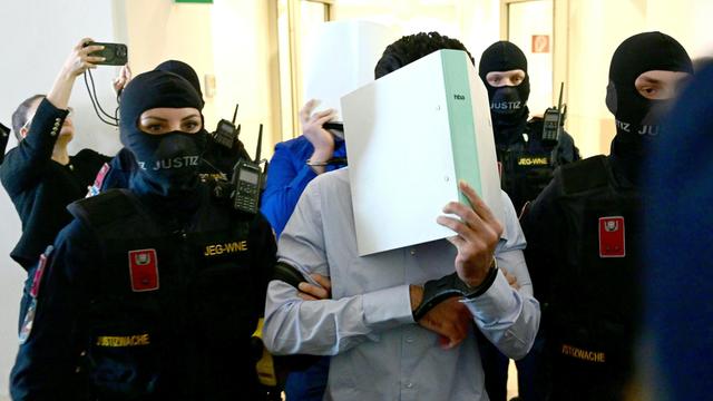 Polizisten führen zwei Männer in in einen Gerichts-Saal. Die Männer tragen Handschellen. Sie halten sich Aktenordner vor das Gesicht.