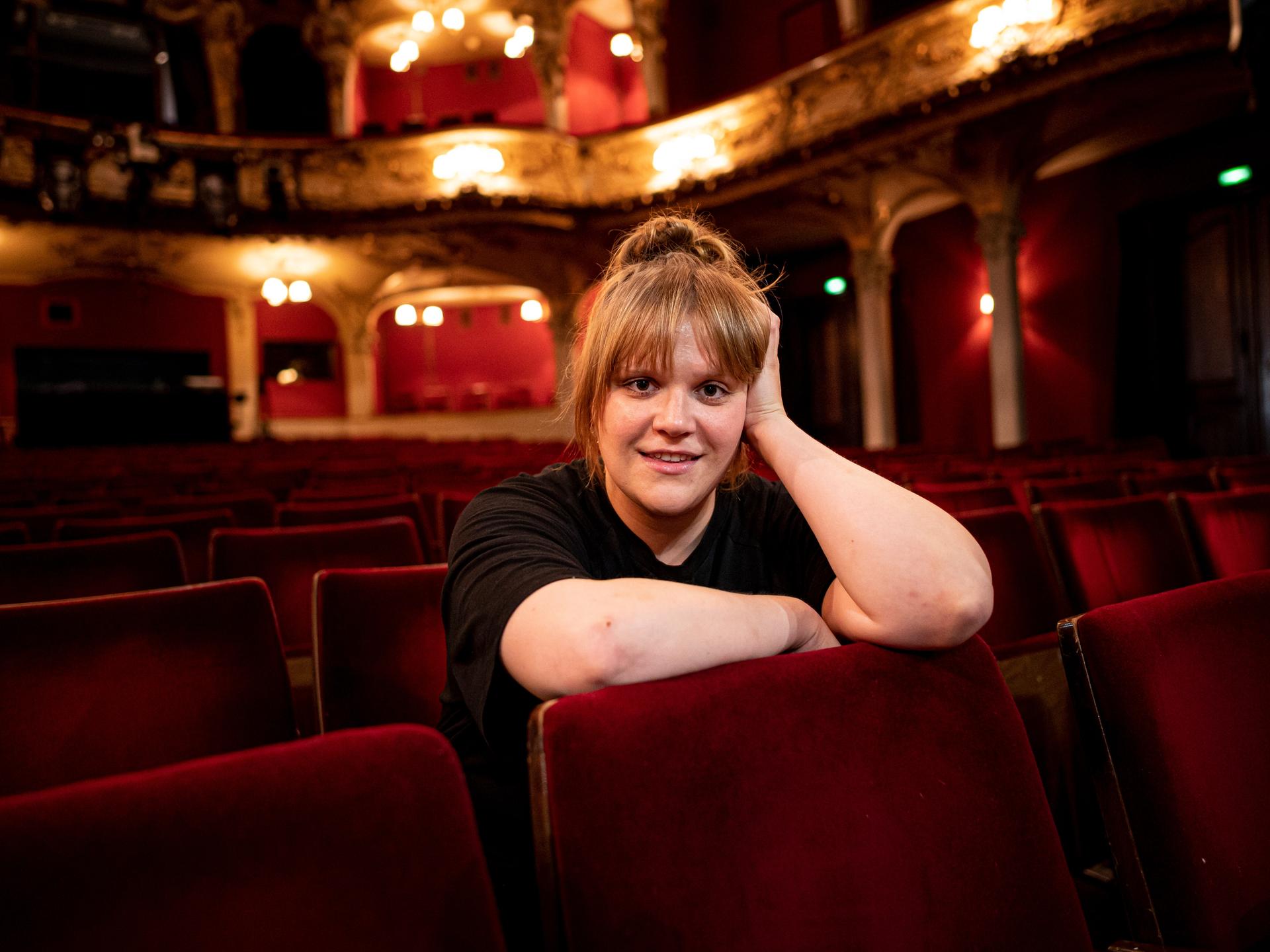Stefanie Reinsperger sitzt im Zuschauerraum des Berliner Ensembles. Sie trägt ein schwarzes T-Shirt und das rotblonde Haar zum Dutt aufgesteckt.