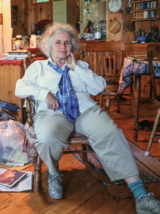 Grace Paley, eine ältere Frau mit grauen Locken, dreckigen Sneakers und einem blauen Halstuch, sitzt in ihrem Zuhause in einem hölzernen Schaukelstuhl, umgeben von Büchern, Pflanzen und Kartons voller Zeitschriften. 