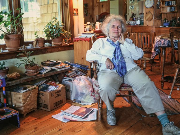Grace Paley, eine ältere Frau mit grauen Locken, dreckigen Sneakers und einem blauen Halstuch, sitzt in ihrem Zuhause in einem hölzernen Schaukelstuhl, umgeben von Büchern, Pflanzen und Kartons voller Zeitschriften. 