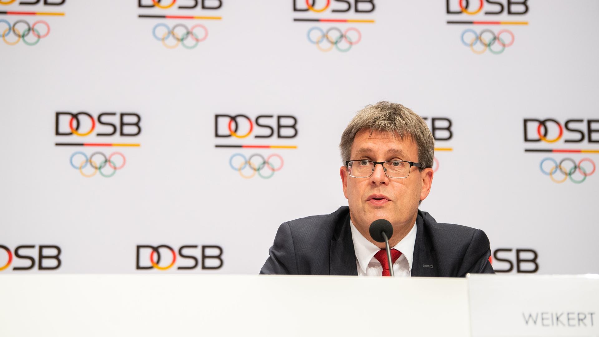 Thomas Weikert, Präsident des Deutschen Olympischen Sportbundes, sitzt auf einer Pressekonferenz vor einer weißen Wand voller  DOSB-Logos und schaut nach vorne.