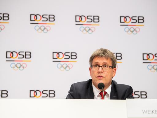 Thomas Weikert, Präsident des Deutschen Olympischen Sportbundes, sitzt auf einer Pressekonferenz vor einer weißen Wand voller  DOSB-Logos und schaut nach vorne.