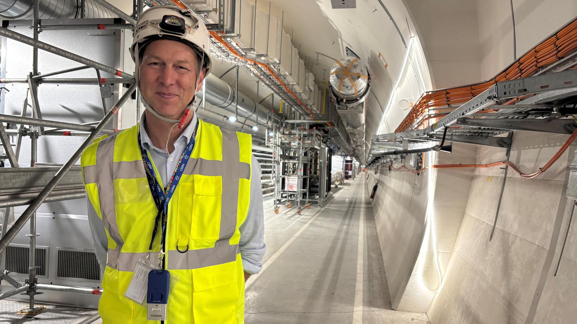 Markus Zerlauth, Projektleiter HL-LHC, steht mit Warnweste und Helm bekleidet in einem neuen Servicetunnel aus Beton mit Kabeln und Rohren.