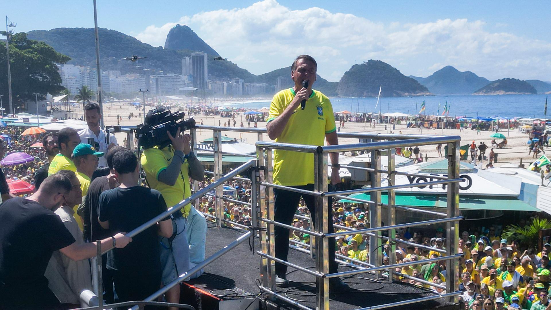 Brasiliens ehemaliger Präsident Jair Bolsonaro steht im gelben T-Shirt auf einem Podest vor Anhängern, die überwiegend gelbe Kleidung tragen. Im Hintergrund sind der berühmte Strand Copacabana sowie die Kulisse des Zuckerbergs zu erkennen.