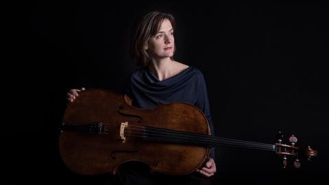 Eine Frau sitzt in dunkler Kleidung vor einem schwarzen Hintergrund. Sie blickt nachdenklich zur linken Seite. Über ihre Beine liegt quer ein Violoncello, das sie mit ihren Händen locker umfasst.