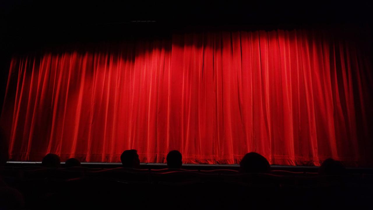 Menschen sitzen in einem Theater vor einem geschlossenen, roten Vorhang. 