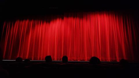Menschen sitzen in einem Theater vor einem geschlossenen, roten Vorhang. 