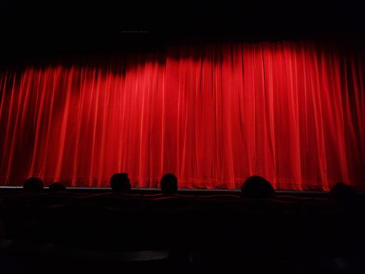 Menschen sitzen in einem Theater vor einem geschlossenen, roten Vorhang. 