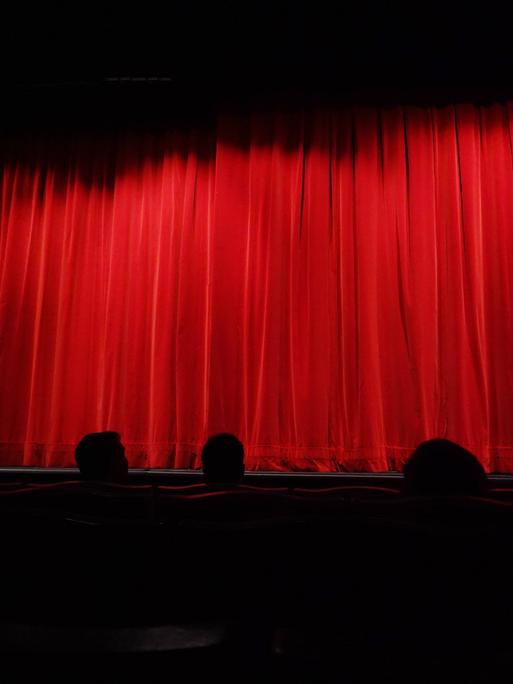 Menschen sitzen in einem Theater vor einem geschlossenen, roten Vorhang. 