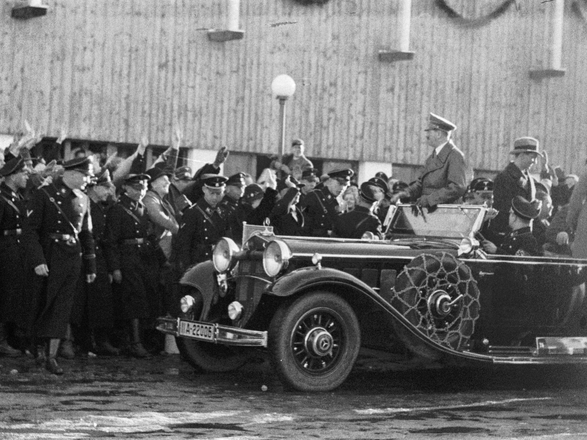 Adolf Hitler wird in einem Auto gefahren, er steht in dem offenen Modell, am Straßenrand jubeln ihm dircht gedrängt Menschen zu. In der ersten Reihe am Straßenrand stehen Uniformierte.