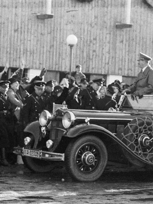 Adolf Hitler wird in einem Auto gefahren, er steht in dem offenen Modell, am Straßenrand jubeln ihm dircht gedrängt Menschen zu. In der ersten Reihe am Straßenrand stehen Uniformierte.