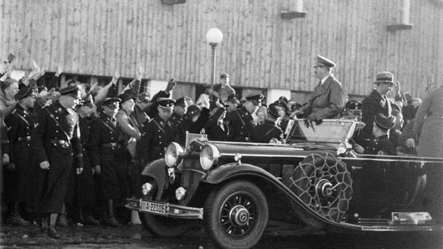 Adolf Hitler wird in einem Auto gefahren, er steht in dem offenen Modell, am Straßenrand jubeln ihm dircht gedrängt Menschen zu. In der ersten Reihe am Straßenrand stehen Uniformierte.