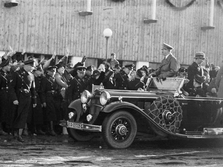 Adolf Hitler wird in einem Auto gefahren, er steht in dem offenen Modell, am Straßenrand jubeln ihm dircht gedrängt Menschen zu. In der ersten Reihe am Straßenrand stehen Uniformierte.